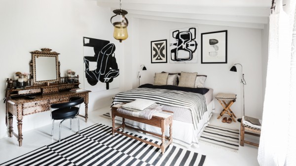 aranżacja sypialni w eklektycznym stylu,dywan w czarno-białe paski,stylowy sekretarzyk,czarno-białe grafiki,stołek drewniany na krzyżaku,eklektyczne wnętrza,dekoracja łóżka w biało-czarnych kolorach,drewniane łóżko,narzuta w paski,