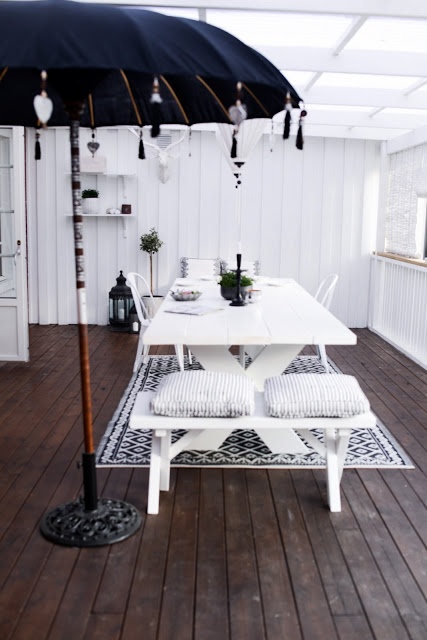 biały drewniany stół na krzyżaku z łąwką,białe meble ogrodowe,czarny parasol tarasowy,jak ozdobić czarny parasol ogrodowy,czarno-biala aranzacja tarasu,biało-czarny dywan w skandynawskim stylu,taras z drewnianych desek,taras w stylu skandynawskim
